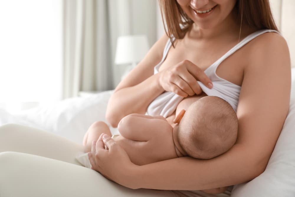 Une femme allaite confortablement son bébé sur un lit, utilisant les éléments indispensables pour un voyage post-partum en douceur et survivre à cette belle phase de la maternité.
